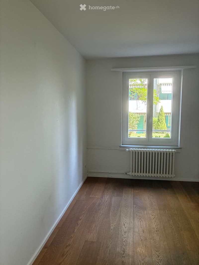 Empty room with wooden flooring, white walls, a window with white frame, and a radiator below the window.