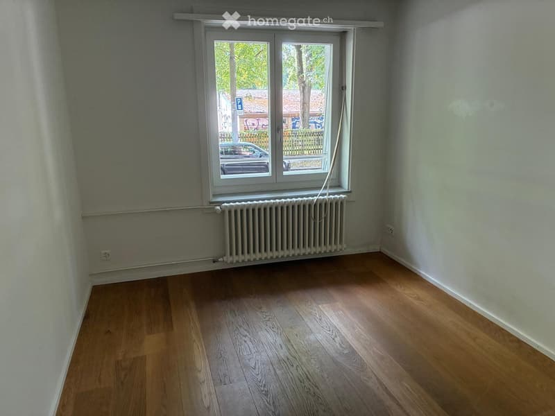 Empty room with wooden flooring, white walls, a radiator, and a large window with a view of the street and parked car.