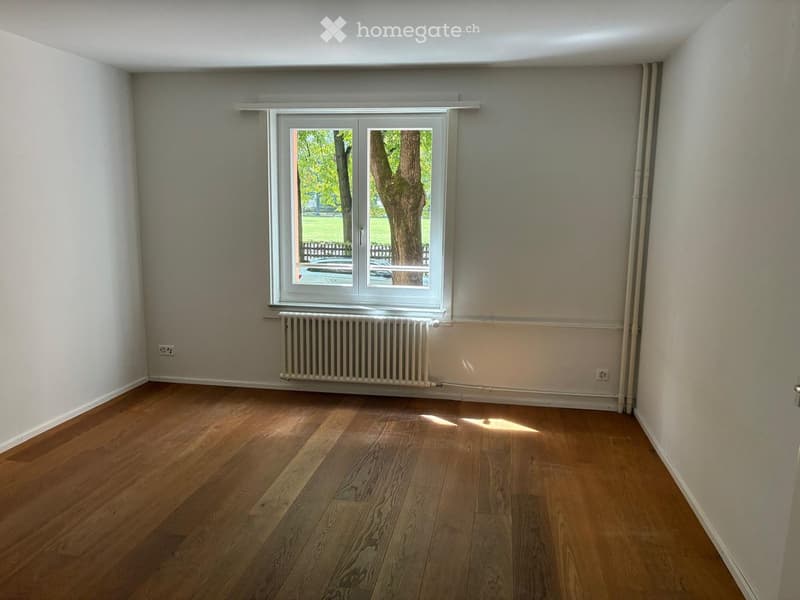 Empty room with wooden flooring, white walls, a window with a view of a tree, and a radiator below the window.
