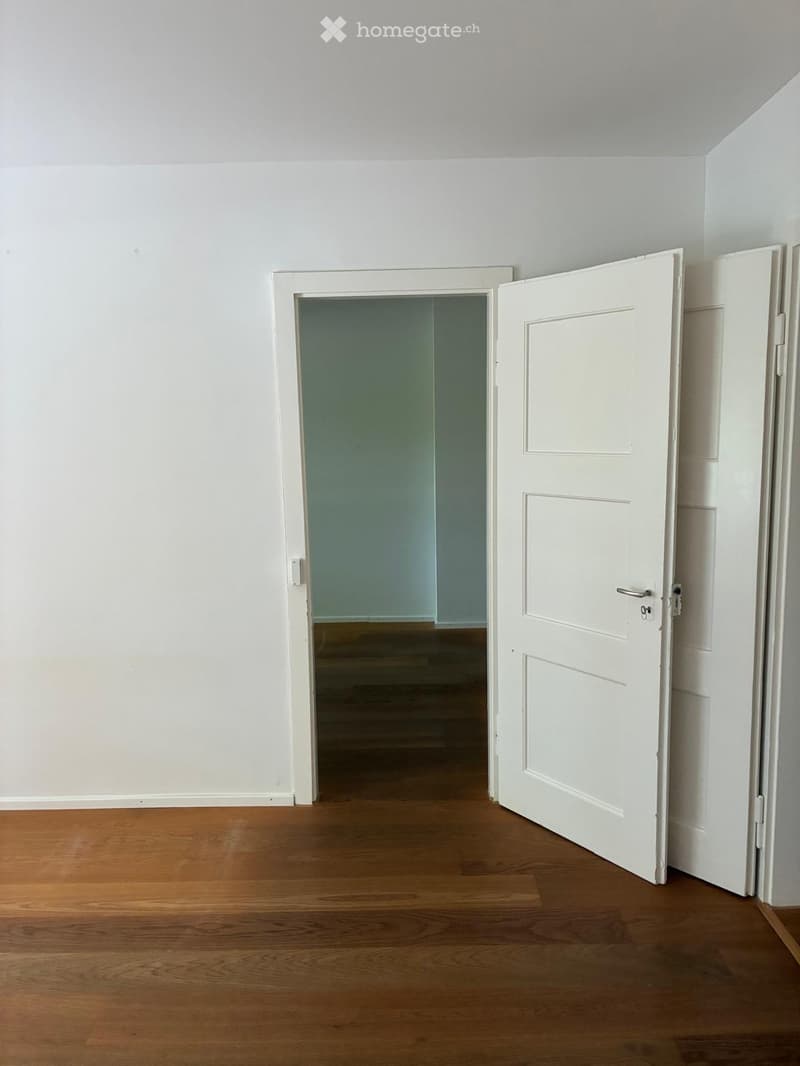 Empty room with wooden flooring and white walls, featuring an open door leading to another room.
