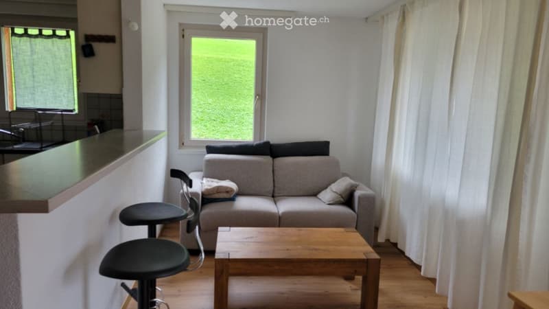 Living room with a grey sofa, wooden coffee table, two black stools, and a window with white curtains.