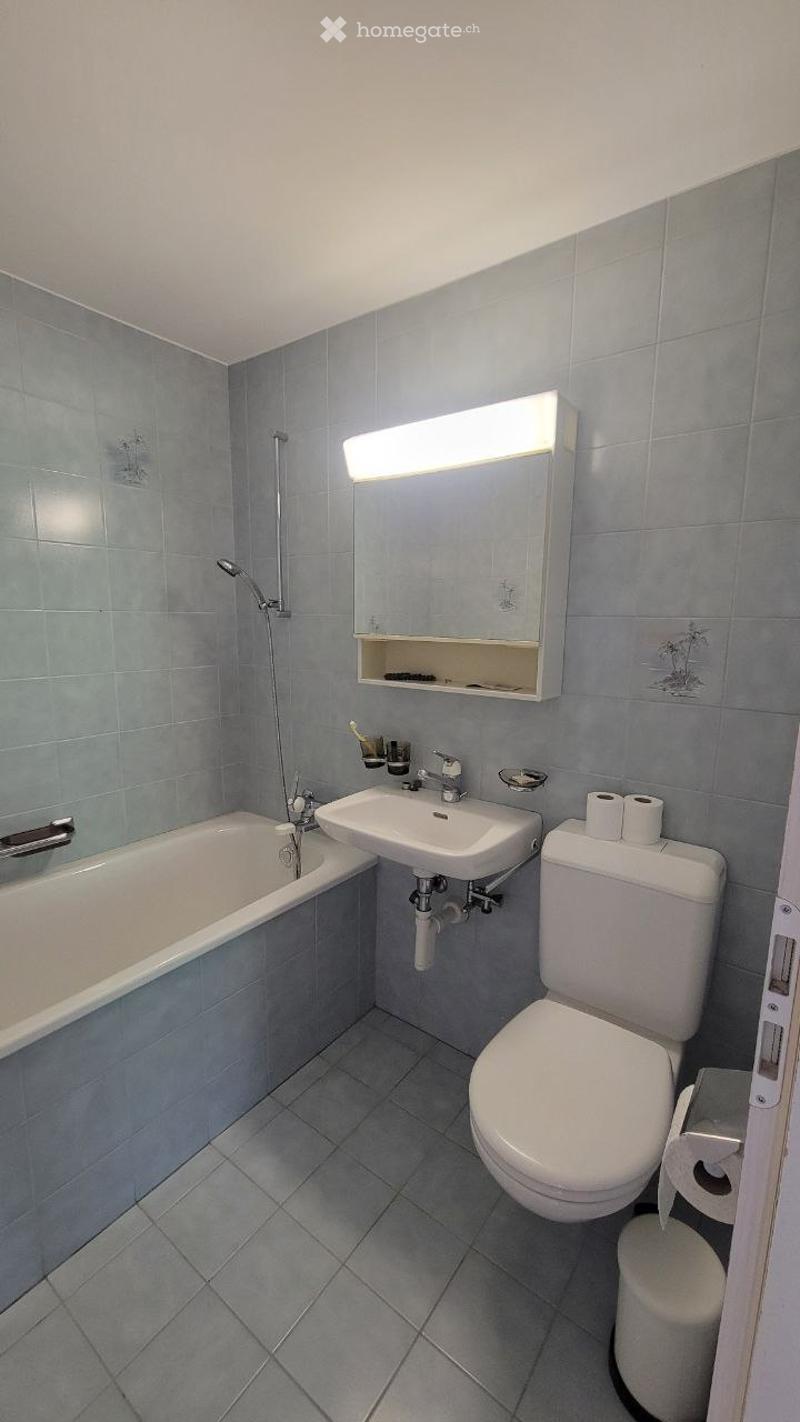 Modern bathroom with a bathtub, sink, toilet, and a mirror with built-in storage. The walls and floor are tiled in a light blue color, and the space is well-lit with a bright light fixture above the mirror.