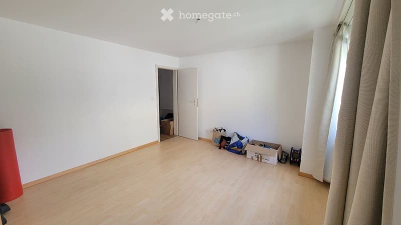 Empty room with light wooden flooring, white walls, and a partially open door leading to another room. Some boxes and items are placed near the door.