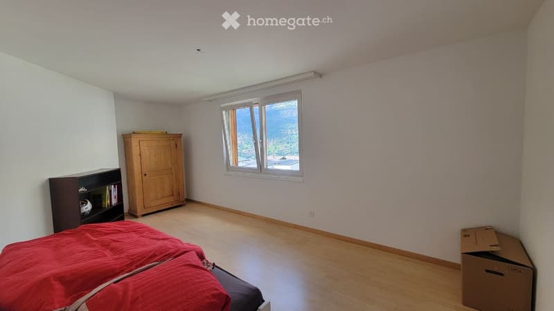 Bright bedroom with wooden flooring, large window offering a scenic view, bed with red bedding, wardrobe, and bookshelf.
