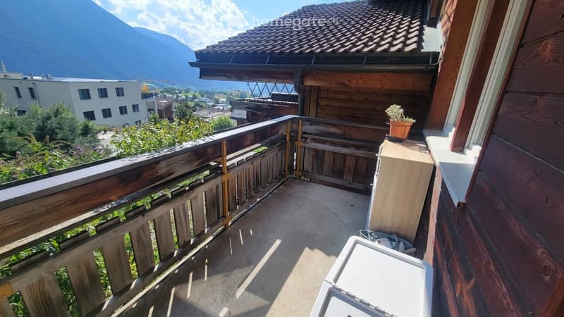Balcony with wooden railing, overlooking a town and mountains, with a small potted plant on the ledge.