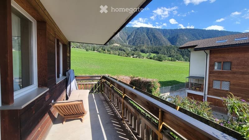 Balcony with wooden railing, overlooking a green valley and mountains, with a wooden chair and small table.