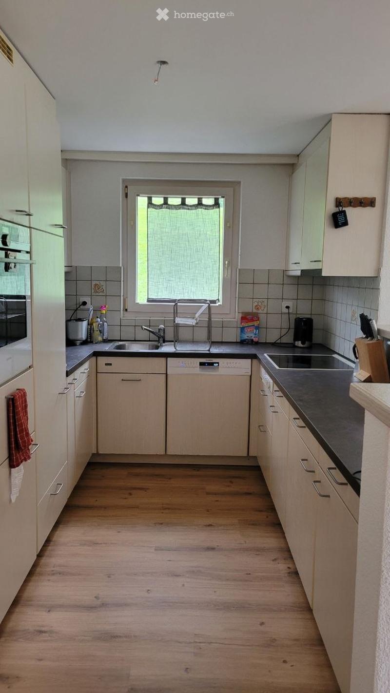 Bright kitchen with light wooden cabinets, stainless steel appliances, and a window above the sink. The floor is wooden, and the countertops are dark grey.