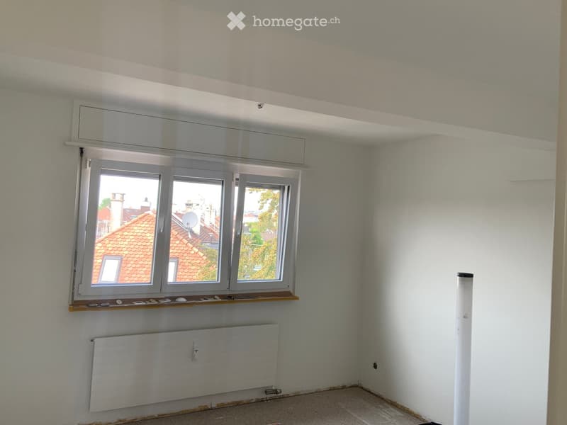 Empty room with white walls, large window with wooden sill, radiator, and visible electrical outlets. No furniture or personal items present.