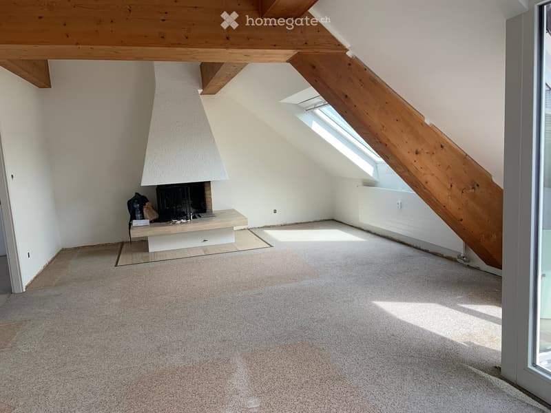 Empty attic space with wooden beams, skylight window, and fireplace area. Bare walls and carpeted floor.