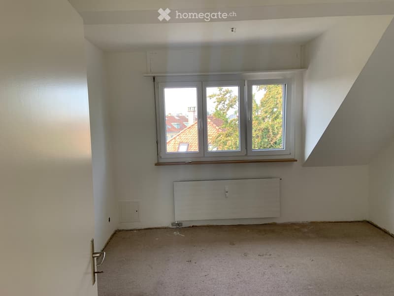Empty room with white walls, a window with a view of rooftops, a radiator, and a door.