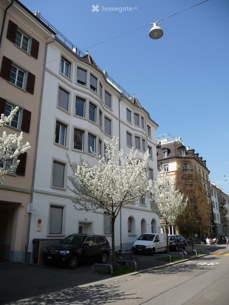 Multi-story white building with numerous windows and balconies, surrounded by blooming trees and parked cars.