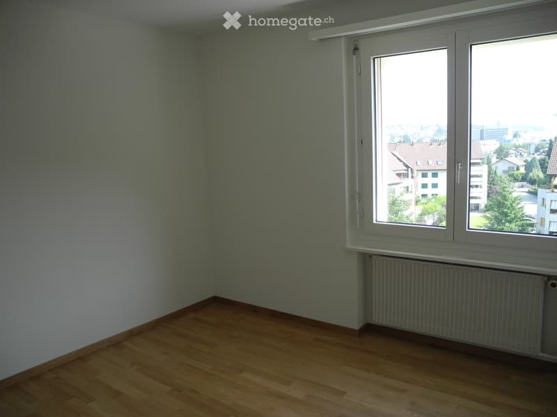 Empty room with wooden flooring, light green walls, and a large window offering a view of the city.