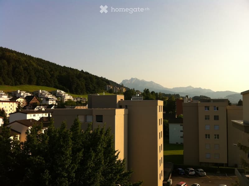 A multi-story residential building with a parking lot in the foreground, surrounded by trees and a scenic mountain backdrop.