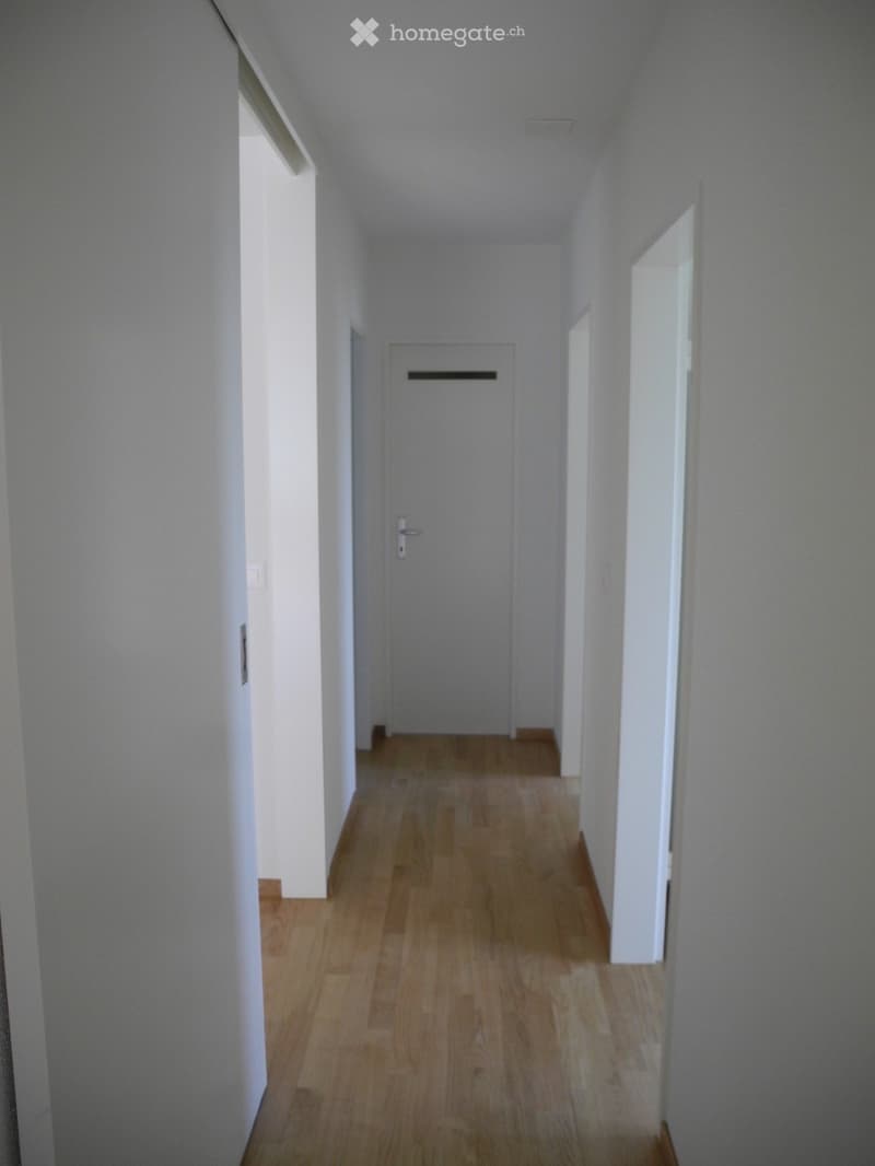 A clean, well-lit hallway with light wooden flooring and white walls, featuring a closed door at the end.