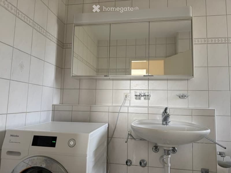 Bathroom with a white sink, mirror cabinet, and a washing machine. The walls are tiled in light grey with a decorative border.