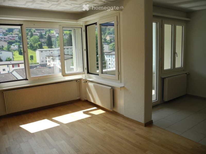 Empty room with wooden flooring, large windows, radiators, and a view of the neighborhood.