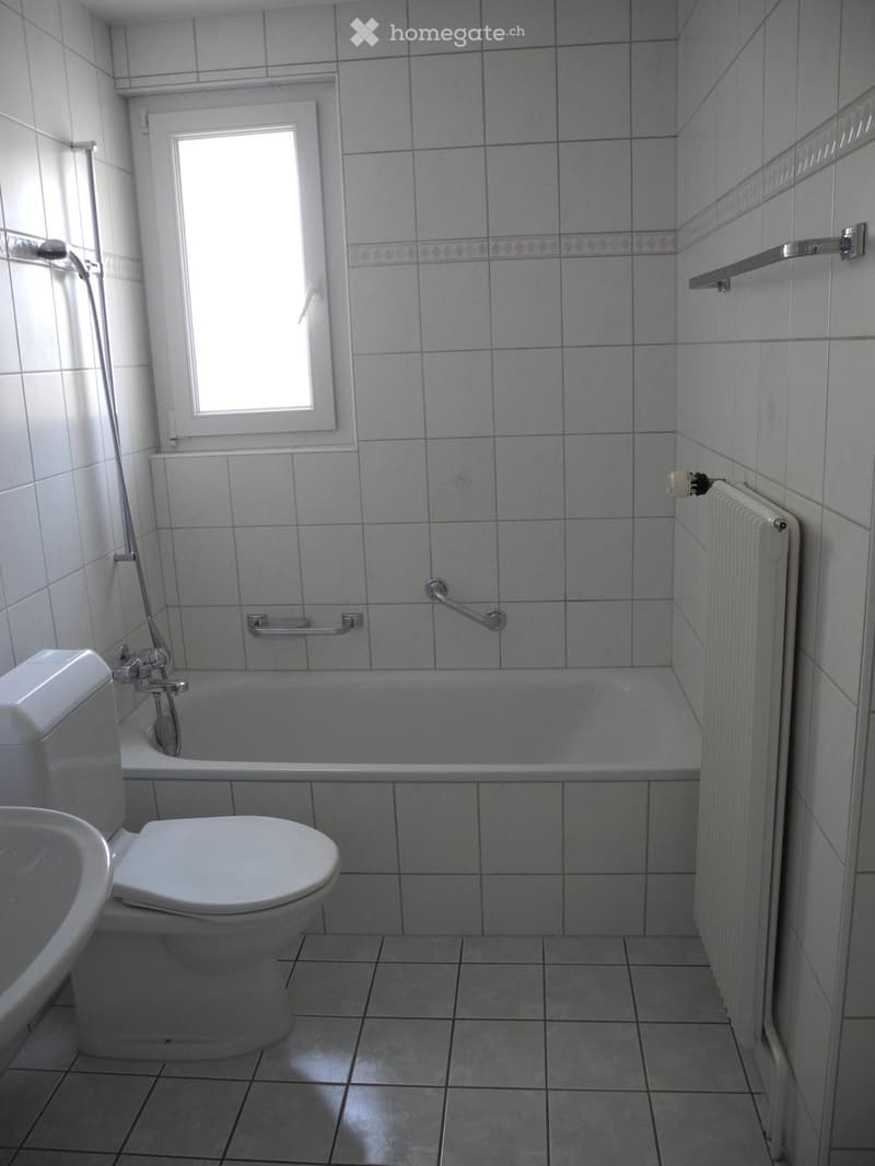 A small bathroom with white tiled walls and floor, featuring a toilet, bathtub, sink, radiator, and handrails for accessibility.