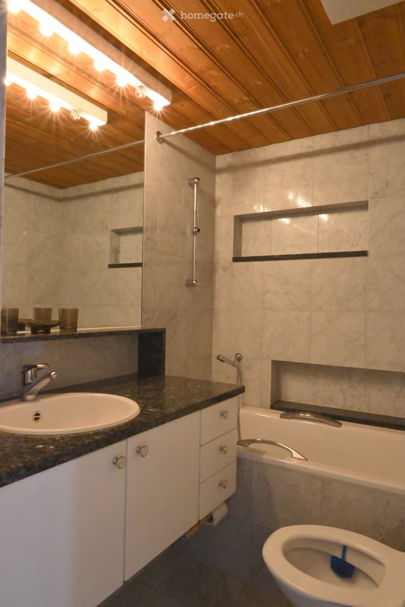 Modern bathroom with a white sink, bathtub, toilet, and mirrored cabinet. Features marble tiles and sleek wooden ceiling with integrated lighting.