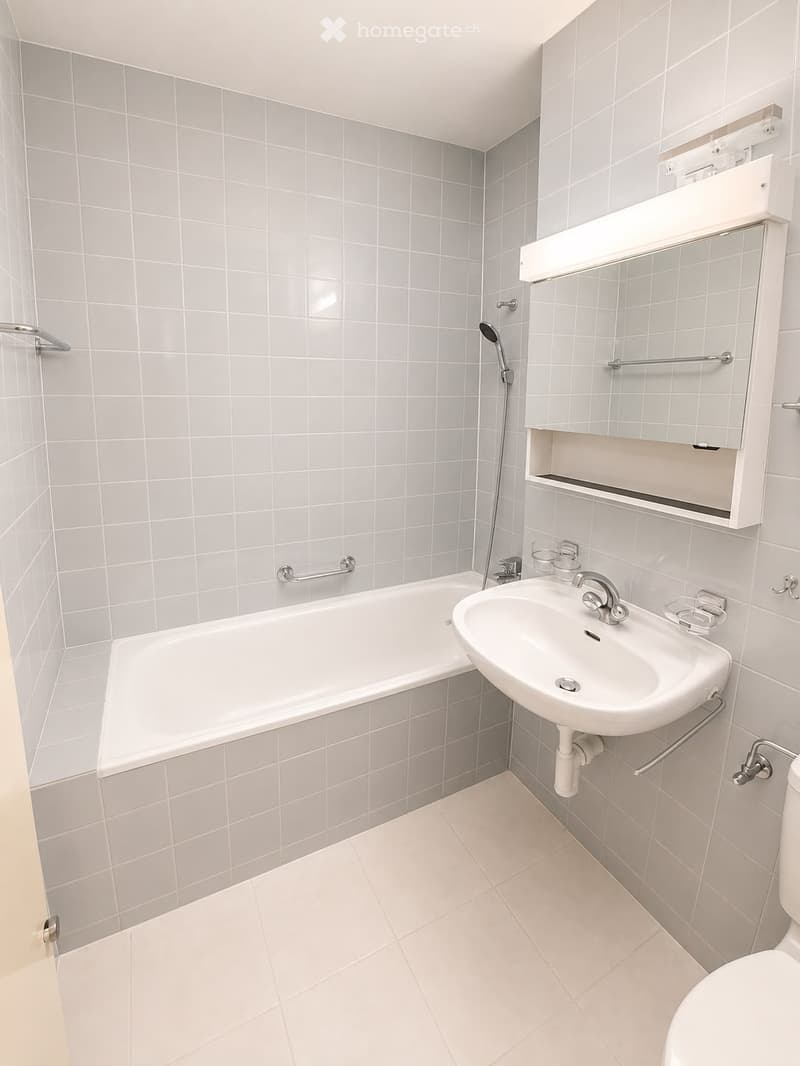 Modern bathroom with white tiled walls and floor, featuring a bathtub, sink, mirror, and toilet.