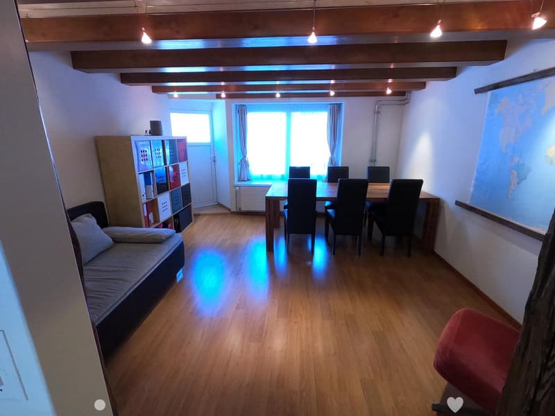 A multi-functional room with a sofa, dining table, chairs, and a storage unit. The room features wooden flooring and large windows with curtains.