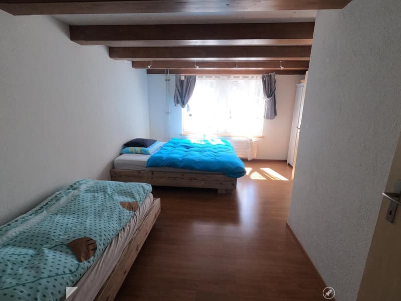 Cozy bedroom with wooden floors, two single beds, and exposed wooden beams on the ceiling. Natural light streams in from a window with curtains.