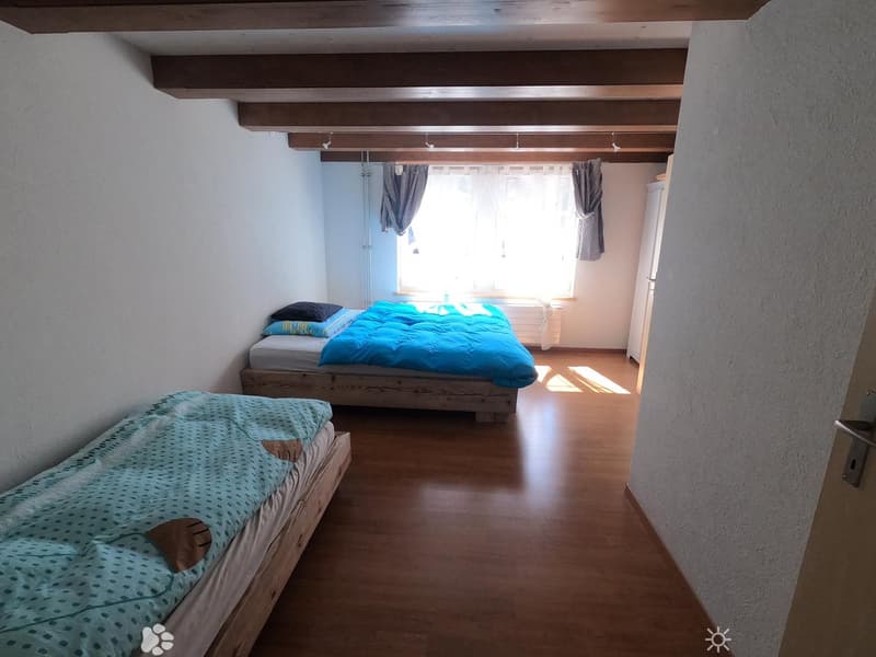 Cozy bedroom with wooden floors, two single beds, and exposed wooden beams on the ceiling. Natural light streams in from a window with curtains.