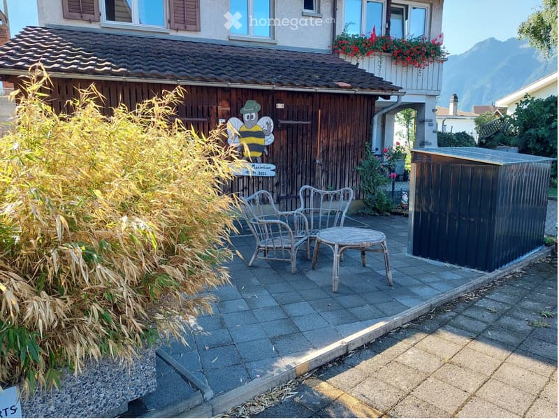 Outdoor patio area with a small table and two chairs, surrounded by tall plants and a wooden fence. A larger house with a balcony and flowers is visible in the background.