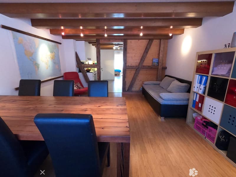 A multi-functional room with a wooden table, chairs, a sofa, and a shelf with storage compartments. The room features exposed wooden beams and a world map on the wall.