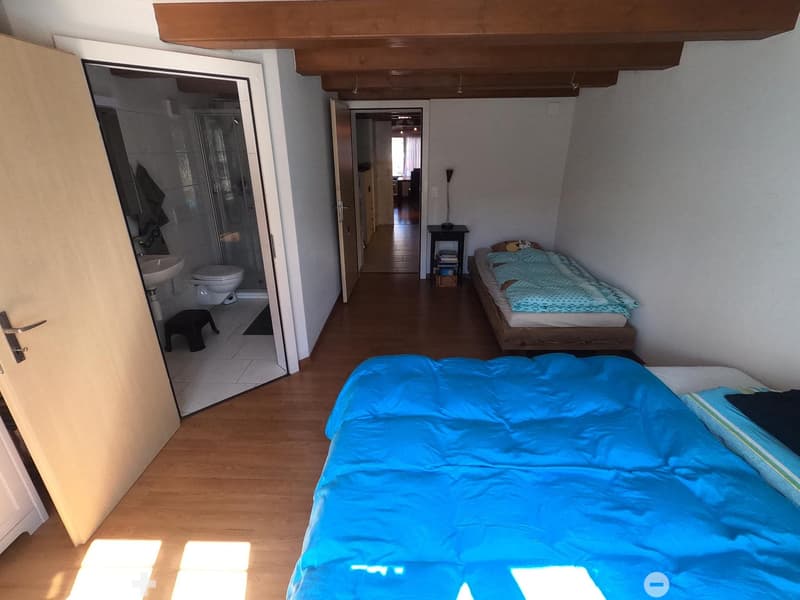 Cozy bedroom with wooden flooring, two beds with blue and green bedding, a small black table with a lamp, a stool, and an open door leading to a bathroom with a sink and toilet.