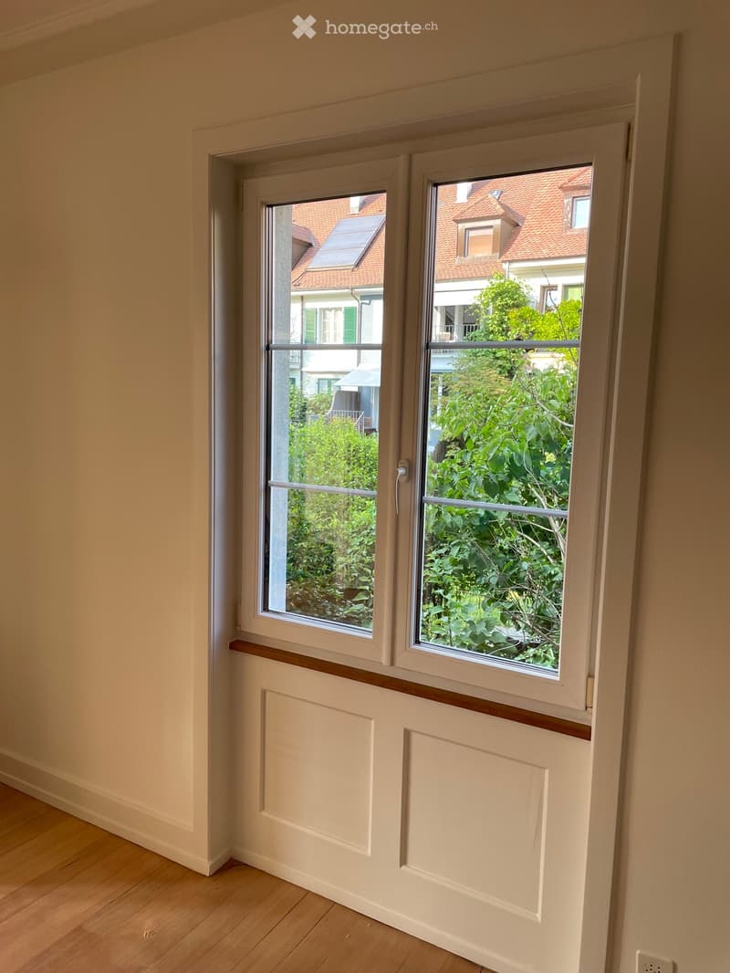 Empty room with light-colored walls, wooden flooring, and two large windows showing a view of neighboring houses and greenery.