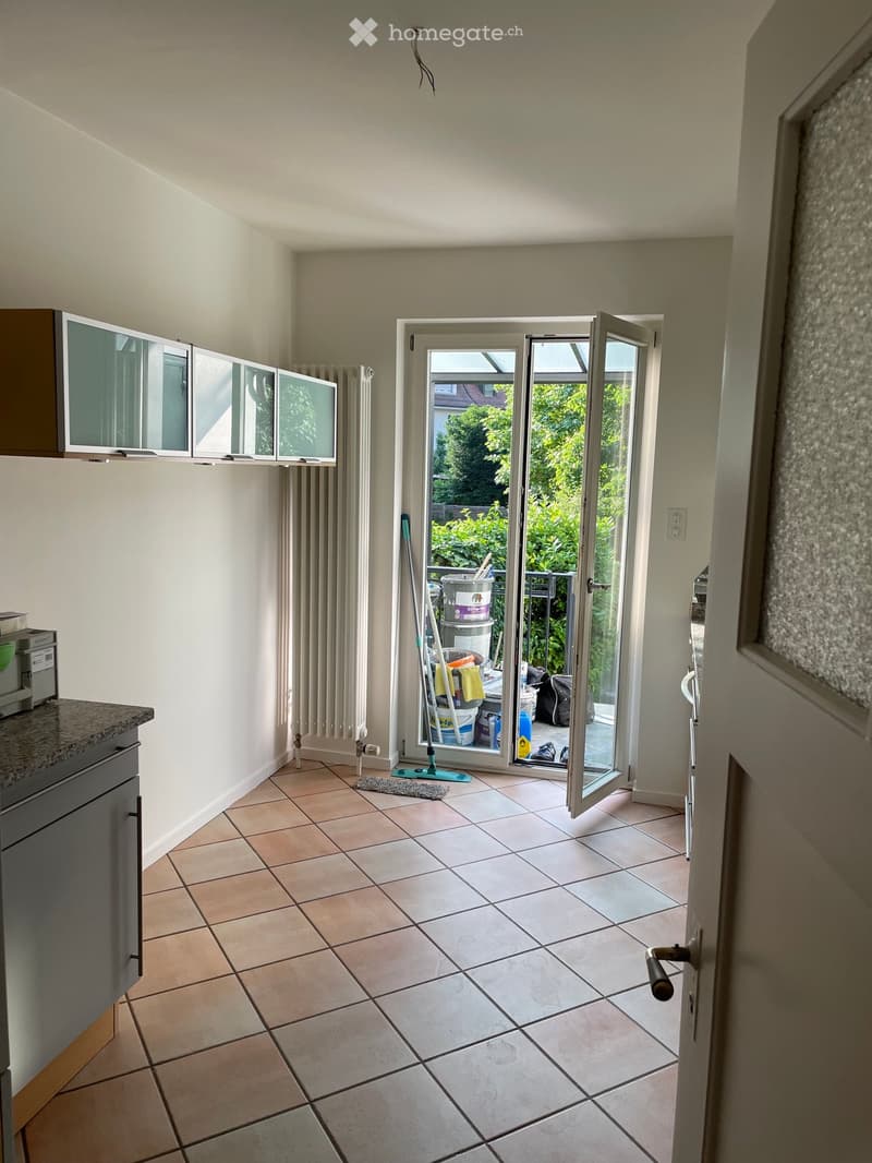 A clean, tiled room with a cabinet, radiator, and a mop leaning against the wall. An open sliding door leads to a balcony with greenery visible outside.