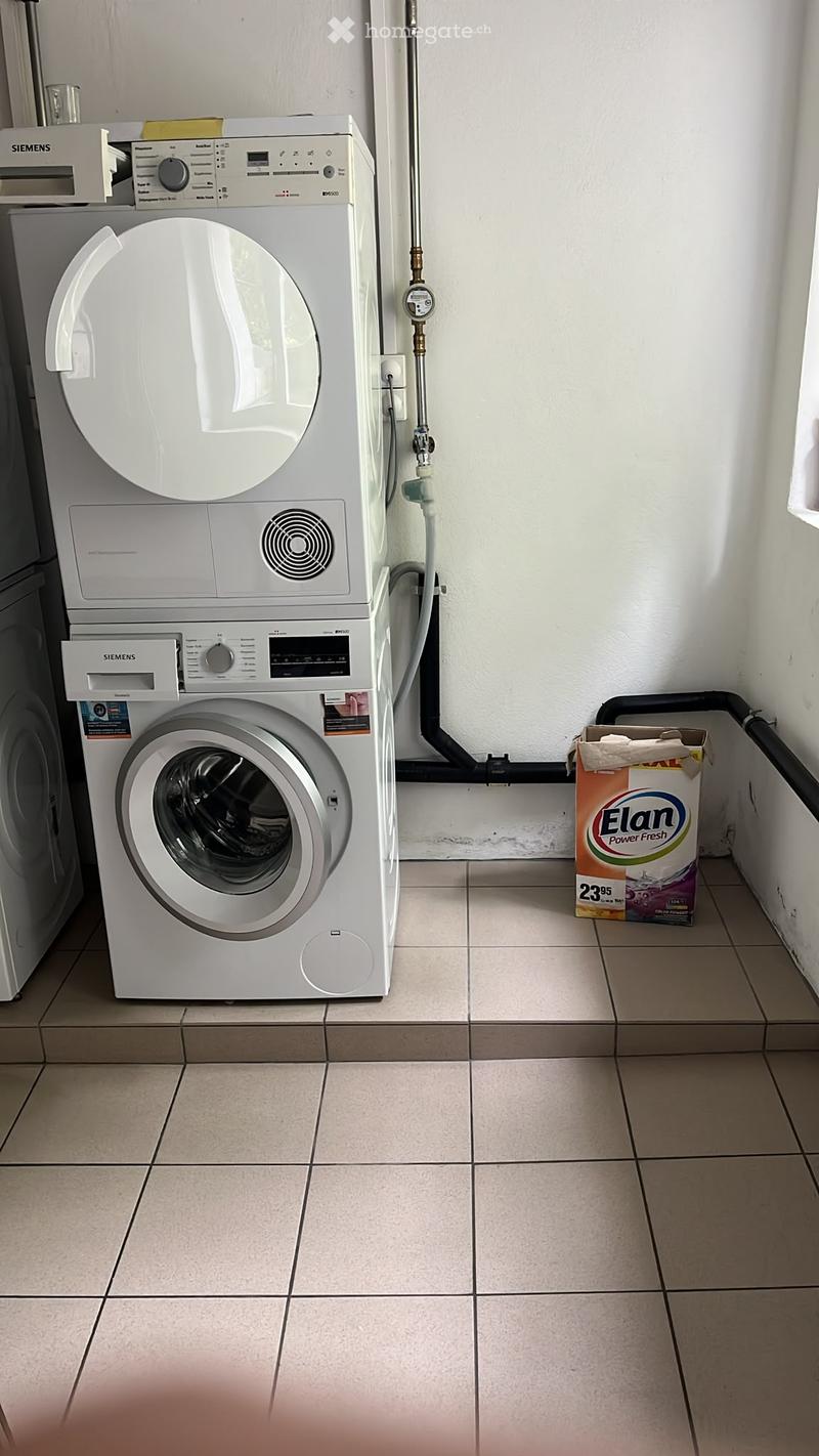 A small bathroom with a washing machine and a dishwasher stacked vertically, tiled floor, and a box of laundry detergent on the floor.