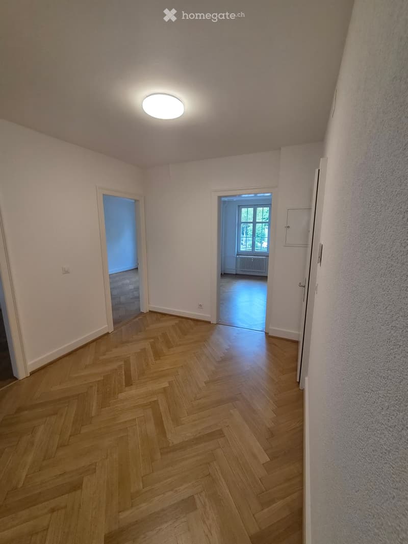 Empty hallway with wooden herringbone flooring, white walls, and recessed lighting. Two doors lead to adjacent rooms, one with a visible window and radiator.