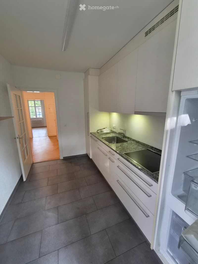 Modern kitchen with white cabinets, granite countertop, sink, and refrigerator. Open doorway leads to another room with wooden flooring and large windows.