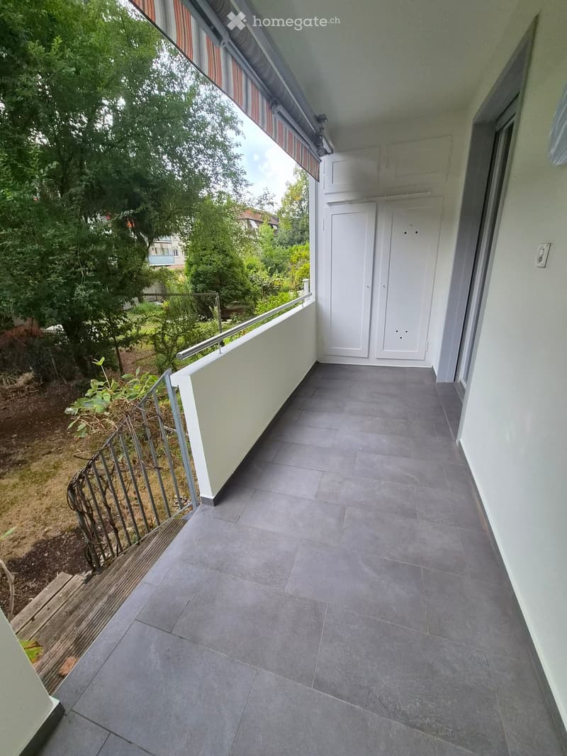 Balcony with grey tiled floor, white double doors, metal railing, and view of greenery.