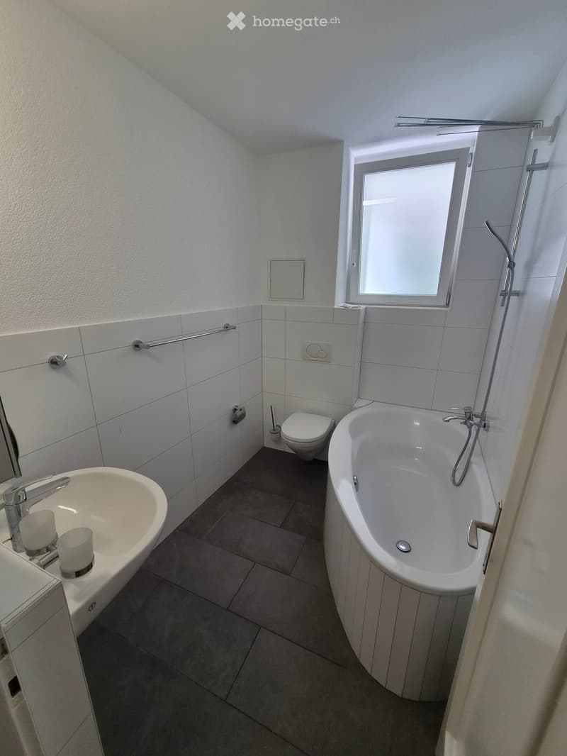 Modern bathroom with a bathtub, toilet, sink, and a window providing natural light. The walls are tiled, and there is a handrail for safety.