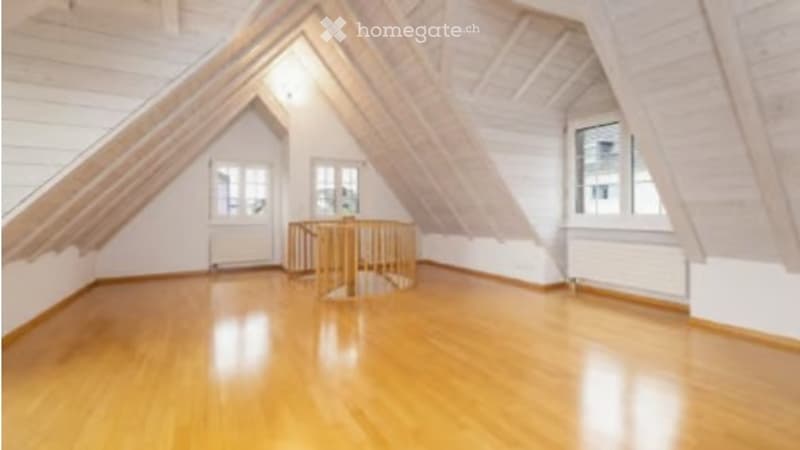 Empty attic space with wooden floors, white walls, and exposed wooden beams. Features a small staircase leading to an upper level and several windows providing natural light.