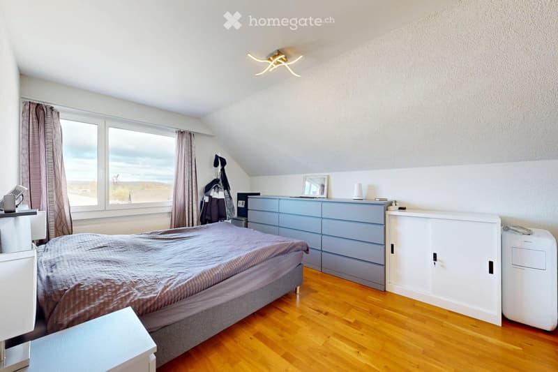 Bright bedroom with wooden flooring, large window offering a scenic view, bed with grey bedding, nightstand, chest of drawers, mirror, and air conditioner.