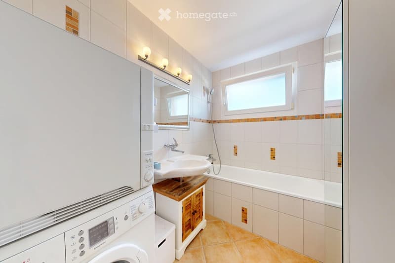 Modern bathroom with white tiles, a sink on a wooden vanity, a bathtub, a mirror, a washing machine, and a window for natural light.