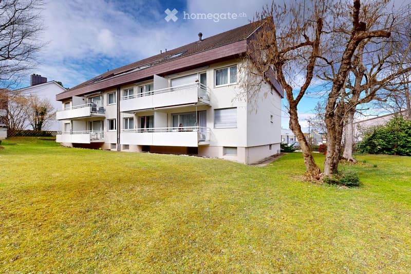 A multi-story residential building with balconies, surrounded by a grassy lawn and trees.