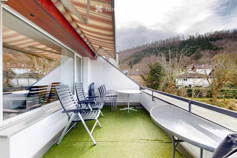 Balcony with green artificial turf, glass railing, and outdoor furniture including a table and chairs, overlooking a hilly landscape.