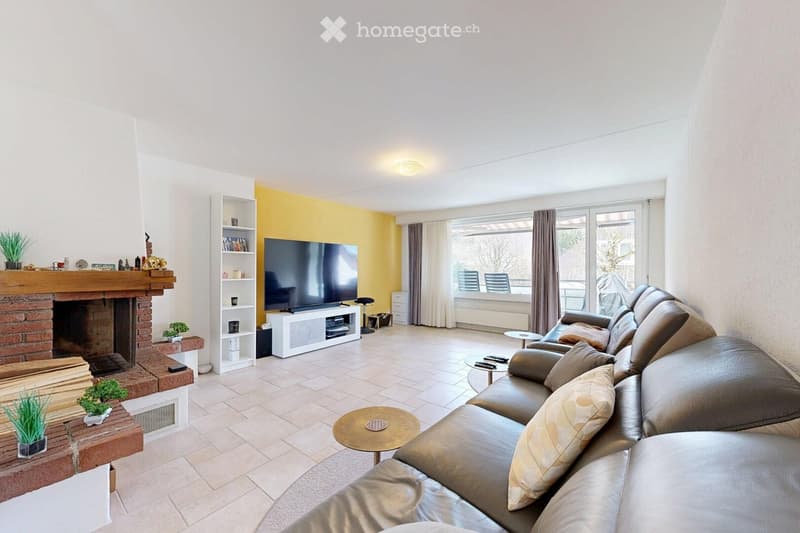 Bright living room with a brick fireplace, large sectional sofa, coffee table, TV, and shelves. Large windows provide natural light.