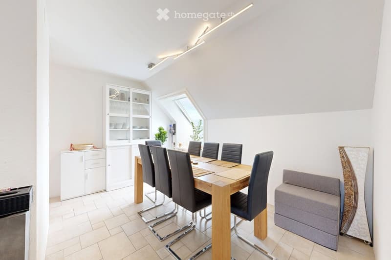Bright dining area with a wooden dining table, six black chairs, a grey sofa, and a white cabinet with glass doors.