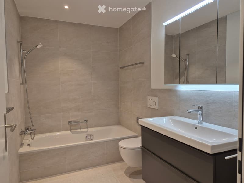 Modern bathroom with beige tiles, a bathtub, toilet, sink, and a large mirror with built-in lighting.