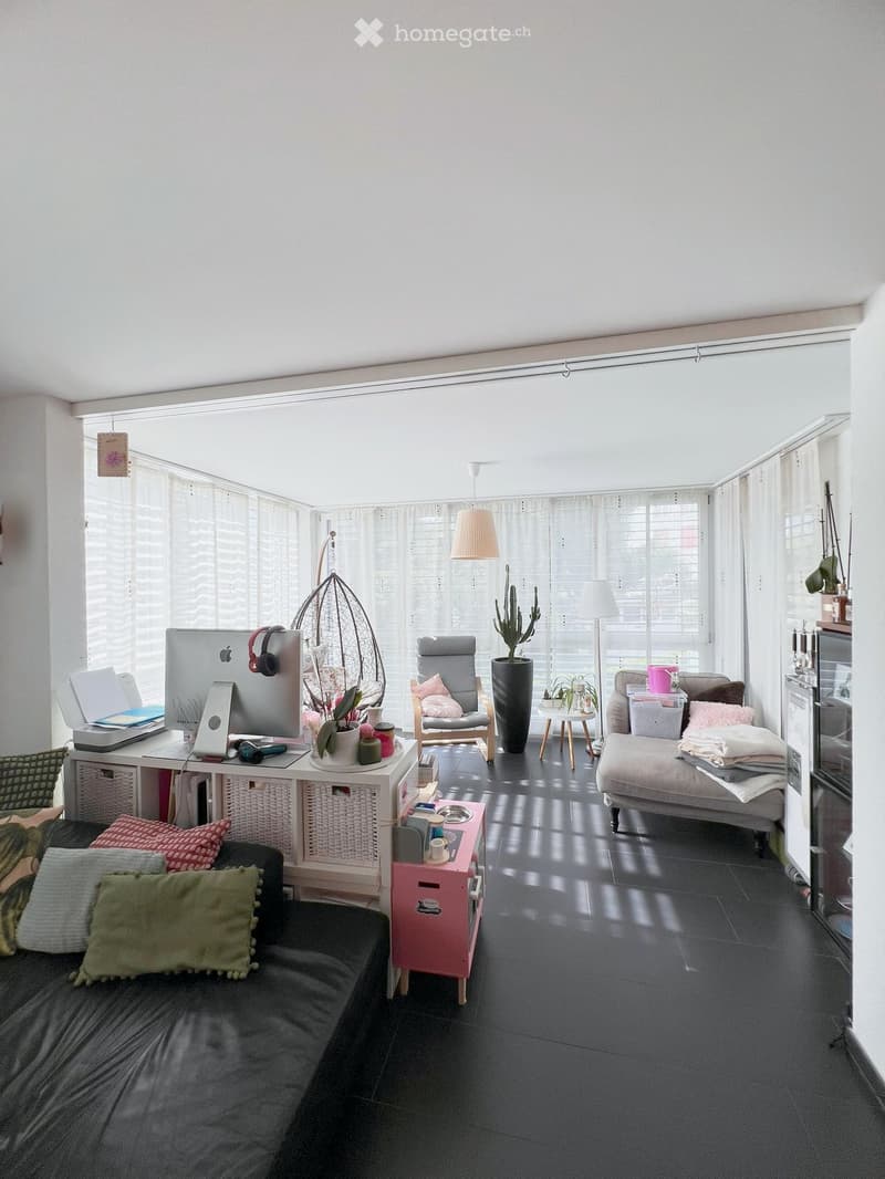 Bright living room with large windows, black sofa, grey armchair, small table, laptop, screen, headphones, decorative plants, and storage cabinet.