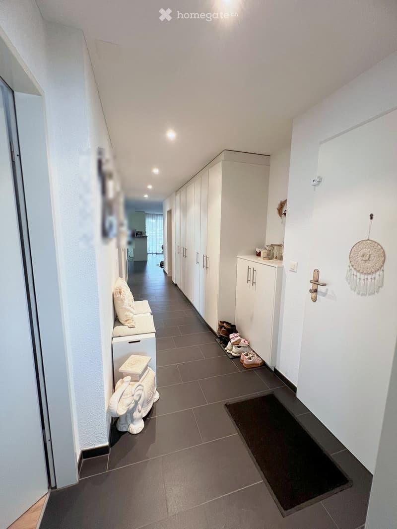 A modern hallway with white walls, dark tiled flooring, built-in white cabinets, and a small seating area with a white bench and decorative elephant sculpture. Shoes are placed near the cabinets, and a dreamcatcher hangs on the wall.