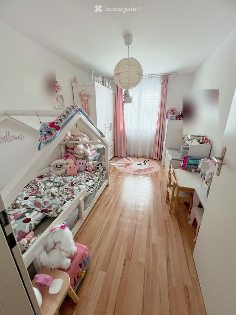 A well-lit bedroom with a wooden floor, a lofted bed with colorful bedding, a desk with a chair, a wardrobe, and decorative items including toys and a round rug.