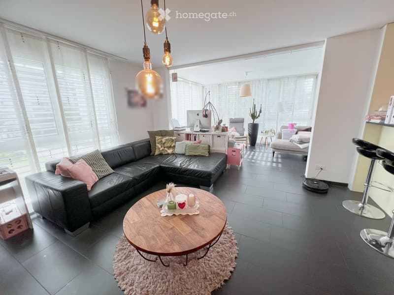 Modern living room with a black sectional sofa, wooden coffee table, hanging pendant lights, and large windows with blinds.