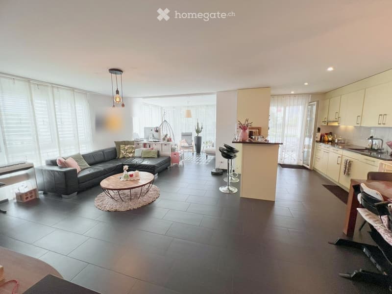 Open-plan living and dining area with a grey sectional sofa, coffee table, dining table, kitchen area with cabinets, and bar stools.