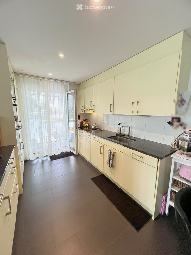 Modern kitchen with white cabinets, dark countertops, and a sink. Features include a built-in oven, a refrigerator, and a small table with a pink container.